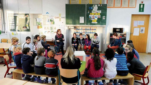 Foto einer Unterrichtssituation: die Lerngruppe sitzt im Halbkreis vor der Tafel. Die Lehrkraft sitzt neben der Tafel.