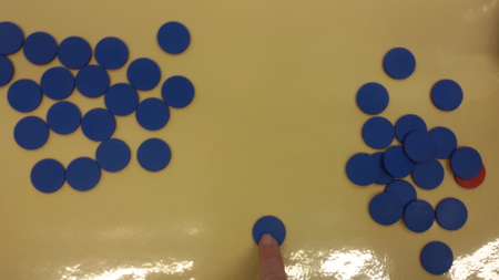 Foto zum Zählen einer Menge unstrukturierter Plättchen. Links unstrukturierter Haufen an blauen Plättchen. Rechts unstrukturierter Haufen blauer Plättchen. Mittig: ein blaues Plättchen auf das ein Finger zeigt.