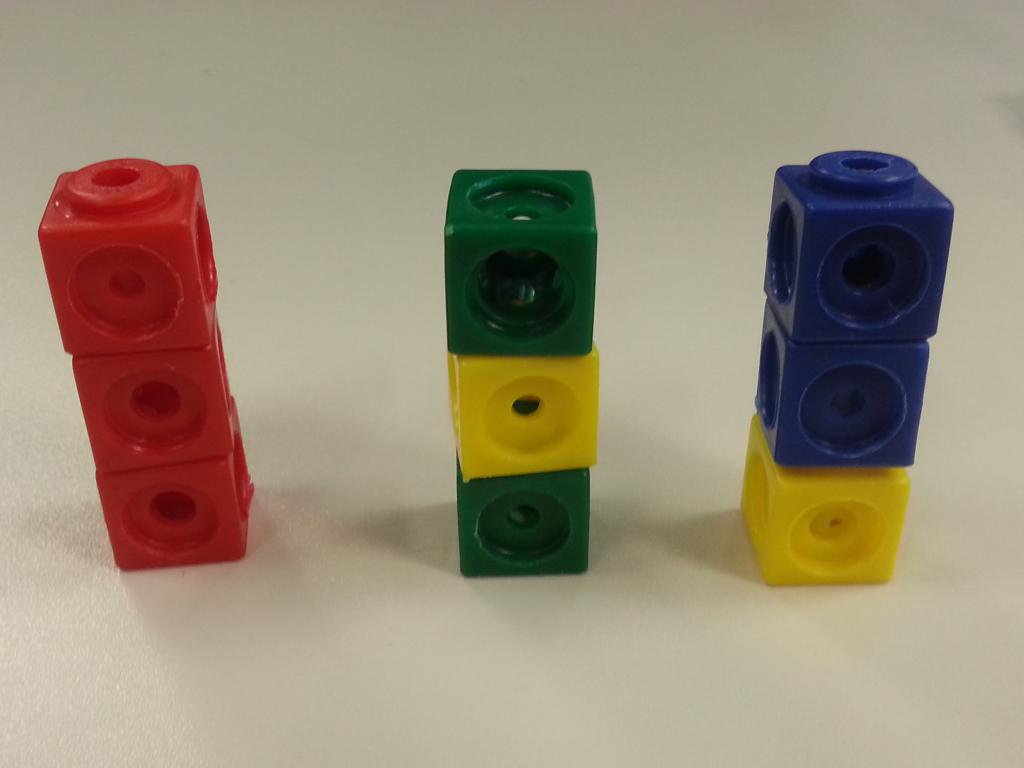 Foto von 3 verschiedenen Steckwürfeltürmen aus jeweils 3 Steckwürfeln:  Links: rot, rot, rot. Mitte: grün, gelb, grün. Rechts: gelb, blau, blau.