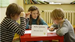 Foto von 3 Kindern, die gemeinsam an einem Tisch sitzen, auf dem ihre Arbeitsblätter liegen. Schild: „Mathe-Konferenz. Bitte nicht stören!“