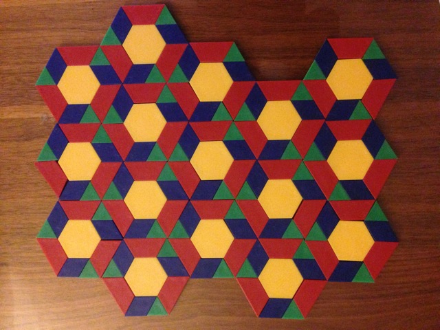 Foto eines Musterparketts, welches im Rahmen des Spiels „Pattern Blocks“ gelegt wurde. Dabei entsteht das Muster durch das Legen einzelner Formen, z. B. Dreieck, Sechseck, Trapez, Parallelogramm.
