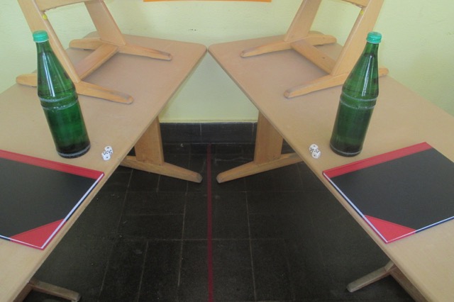 Foto eines Klassenraums, indem 2 Tische symmetrisch angeordnet wurden. Auf diesen befindet sich ebenfalls symmetrisch jeweils eine Wasserflasche, 2 Spielwürfel und eine Kladde. Auf den Boden ist mit Klebeband eine Symmetrieachse geklebt.