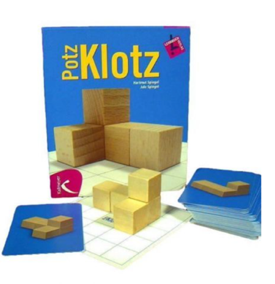Foto des Spiels „Potz Klotz“. Vor dem aufgestellten Spielkarton  liegt ein Stapel mit Spielkarten auf denen Würfelgebäude abgebildet sind und ein gebautes Würfelgebäude auf einer Bauunterlage.
